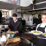 Successful young waitress standing in restaurant kitchen with ordered meals, ready to serving guests